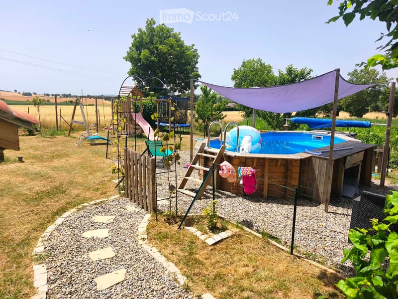 Outdoor area with a small swimming pool, surrounded by gravel and wooden decking, with a canopy providing shade, a slide, and a swing set in the background.