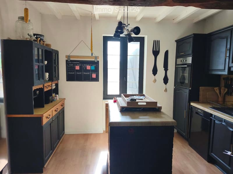 Modern kitchen with dark cabinets, a central island, and various appliances including a refrigerator, oven, and dishwasher. The island features a wooden countertop with a tray and other items. A menu board is visible on the wall, and utensils are displaye