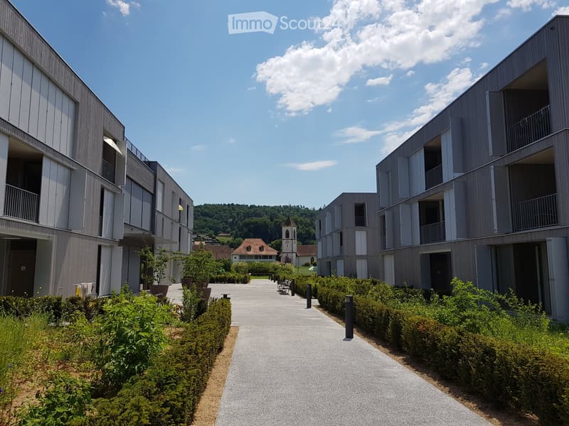 Modern residential complex with grey facades, balconies, and a landscaped courtyard with pathways and greenery.