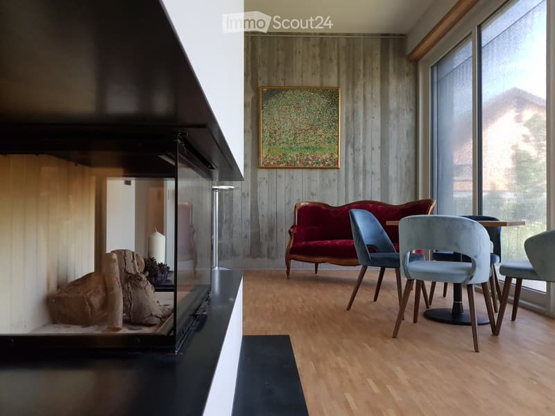 Modern living room with a red velvet sofa, light blue chairs, a small table, a fireplace with logs, and a framed artwork on the wall.