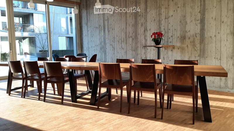 Modern dining area with a large wooden table, multiple chairs, and a vase with red flowers on a side table. Large windows provide natural light.
