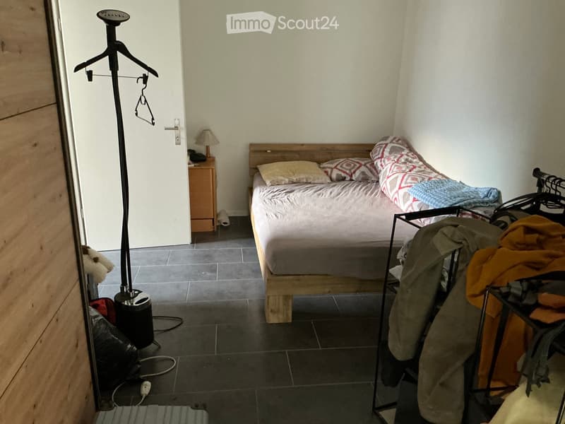 Cozy bedroom with a wooden bed, nightstand, lamp, and clothing rack. The room features tiled flooring and a partially open door leading to another area.