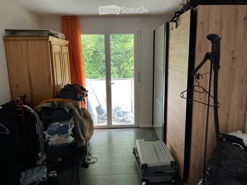 A small, furnished room with a wardrobe, a clothing rack, a suitcase, and a stack of clothes near the sliding glass door leading to a balcony.