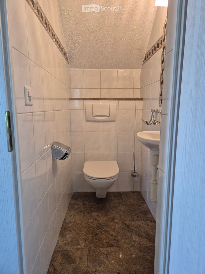 Modern bathroom with white tiled walls, a toilet, a sink with a chrome faucet, and a toilet paper holder. The floor is made of dark brown tiles.