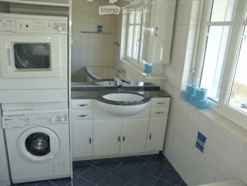 Bathroom with a sink, faucet, mirror, cabinets, and a stacked washing machine and dryer unit. The room features white tiles and a dark blue tiled floor.