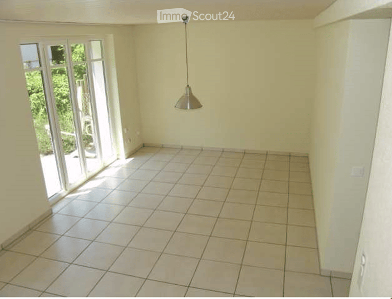 Empty room with tiled floor, white walls, large sliding glass door leading to outdoor area, and a single hanging light fixture.