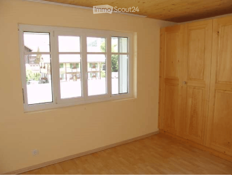 Empty room with light wooden flooring, white walls, and a large window with multiple panes. A built-in wooden closet is visible on one side.