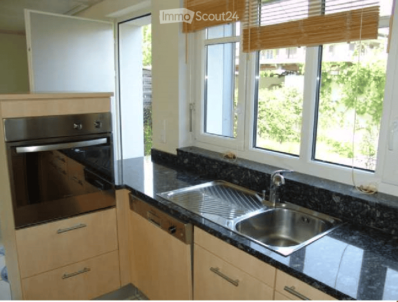 Modern kitchen with light wooden cabinets, stainless steel sink, and built-in oven. Granite countertop and large windows provide natural light.