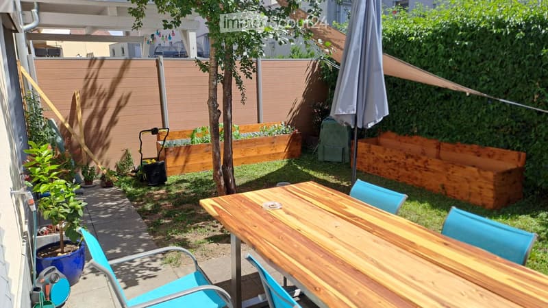 Garden area with wooden raised beds, a small table with chairs, potted plants, and a tree, enclosed by a wooden fence and greenery.