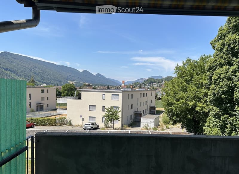View from a balcony showing a multi-story residential building, parked cars, trees, and distant mountains under a clear blue sky.