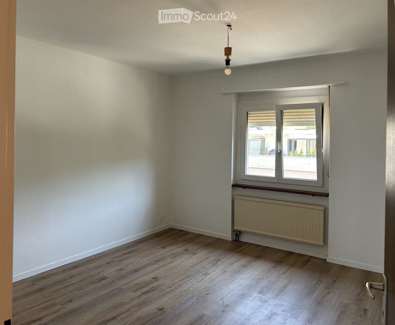 Empty room with light-colored wooden flooring, white walls, a window with blinds, and a radiator below the window. The room has a simple, clean design with no furniture.