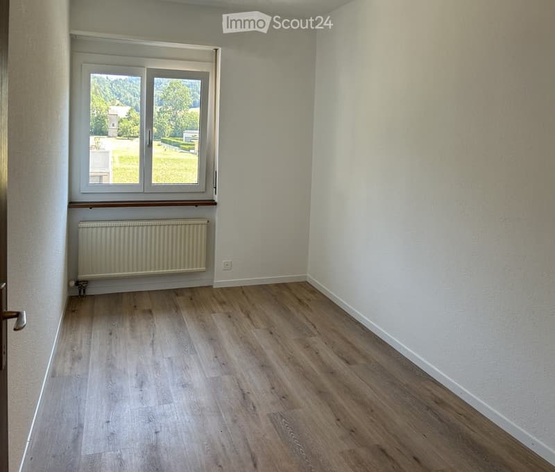 Empty room with light wooden flooring, white walls, a window with a radiator below, and a door on the left side.