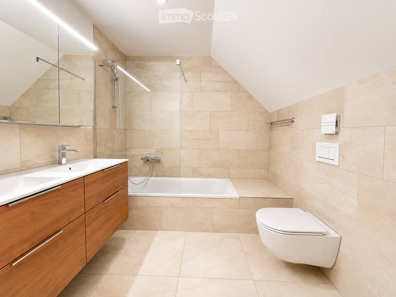 Modern bathroom with a bathtub, toilet, sink, and wooden cabinet. Features a large mirror, sleek fixtures, and warm beige tiles.