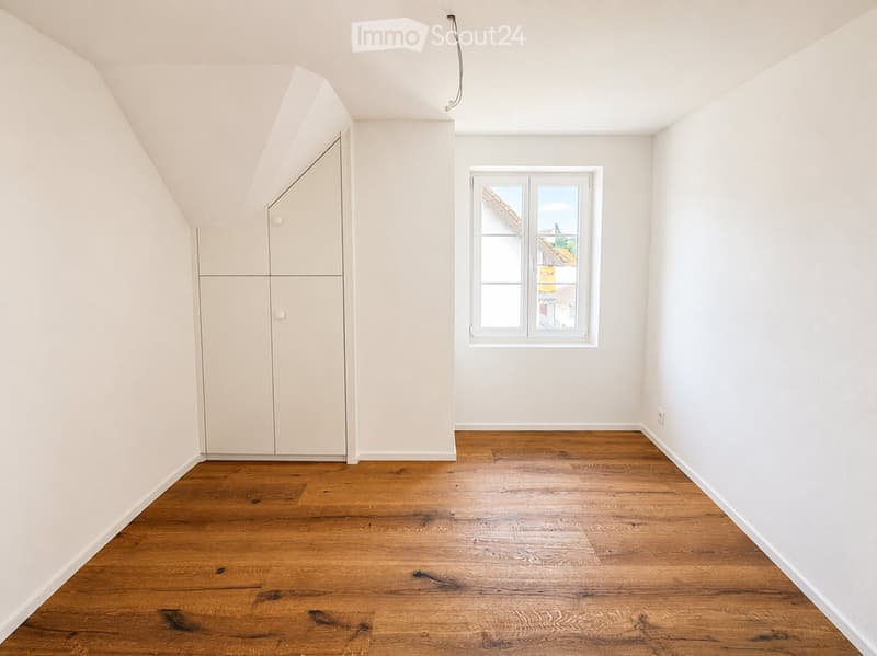 Empty attic room with wooden flooring, white walls, and a built-in closet. A window offers a view of the outside.