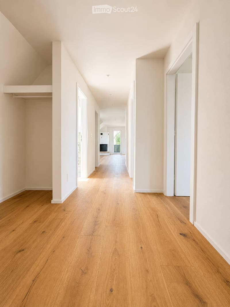 Empty hallway with light wooden flooring and white walls, leading to an open area with a visible TV and greenery in the distance.