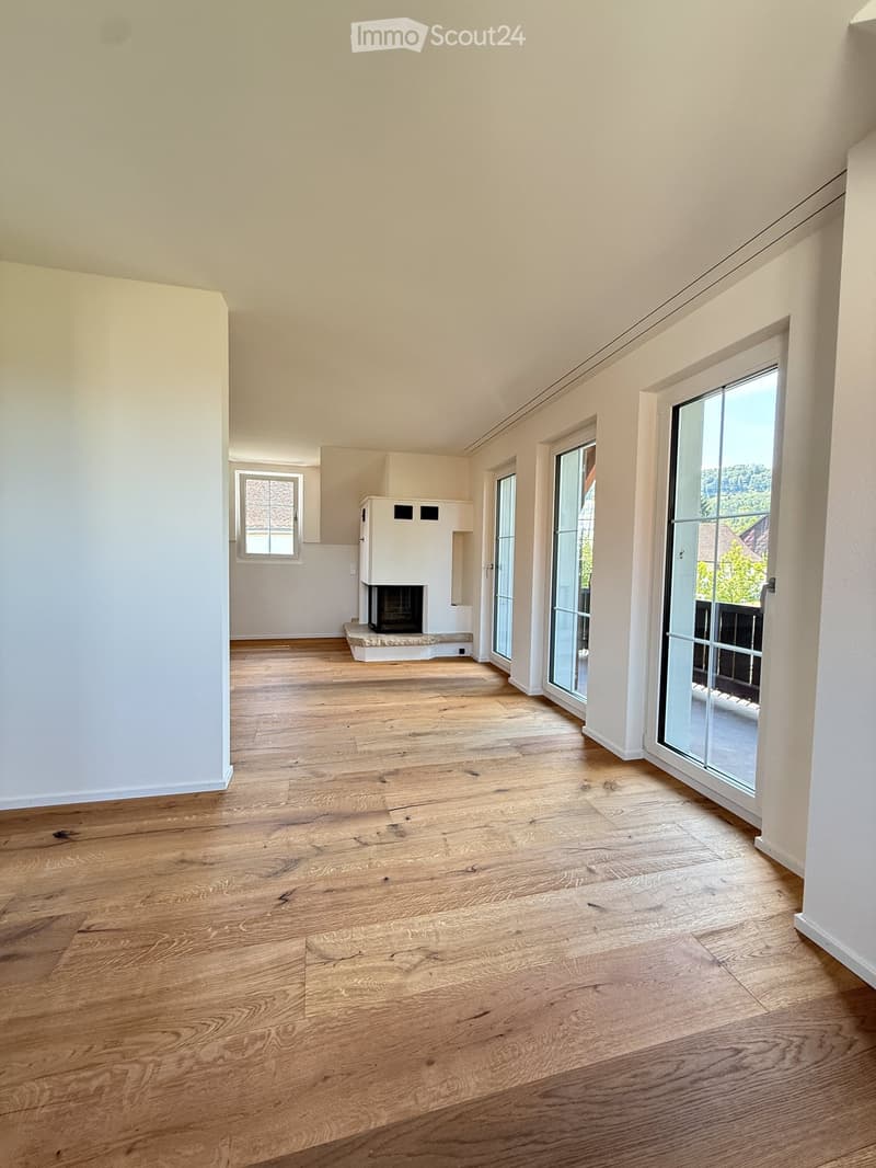 Empty room with light wooden flooring, white walls, and large windows. Features a fireplace and glass doors leading to an outdoor area.