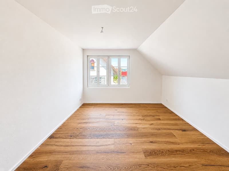 Empty attic room with wooden flooring and white walls, featuring a single window with a view of the outside.