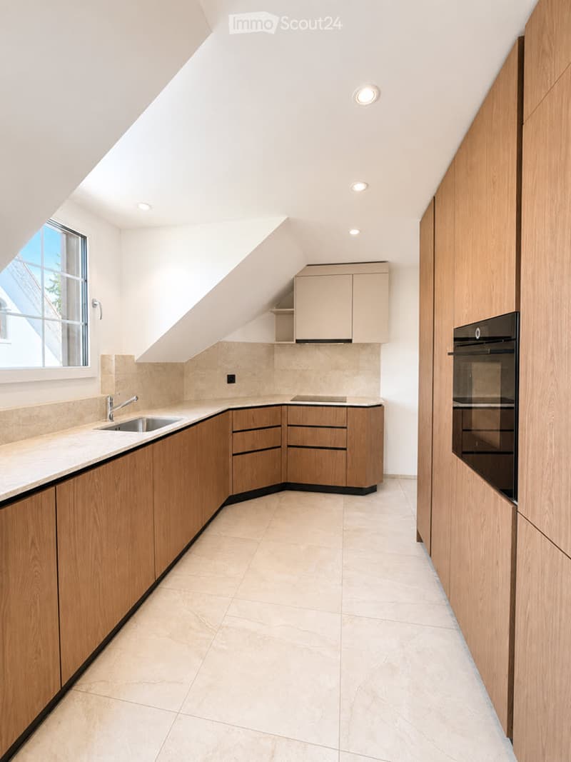Modern kitchen with light wood cabinets, marble countertops, and built-in appliances, featuring a skylight and large window for natural light.