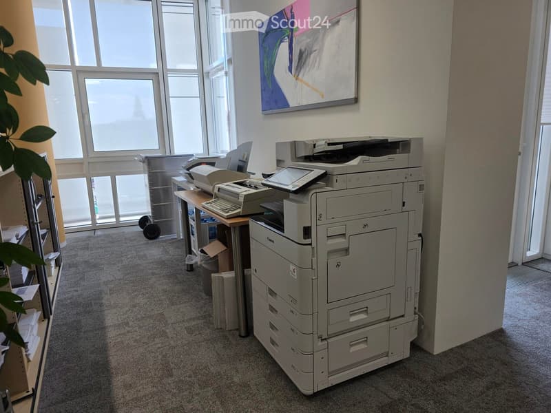 Office space with a large printer, table, keyboard, screen, chair, and plant near large windows.