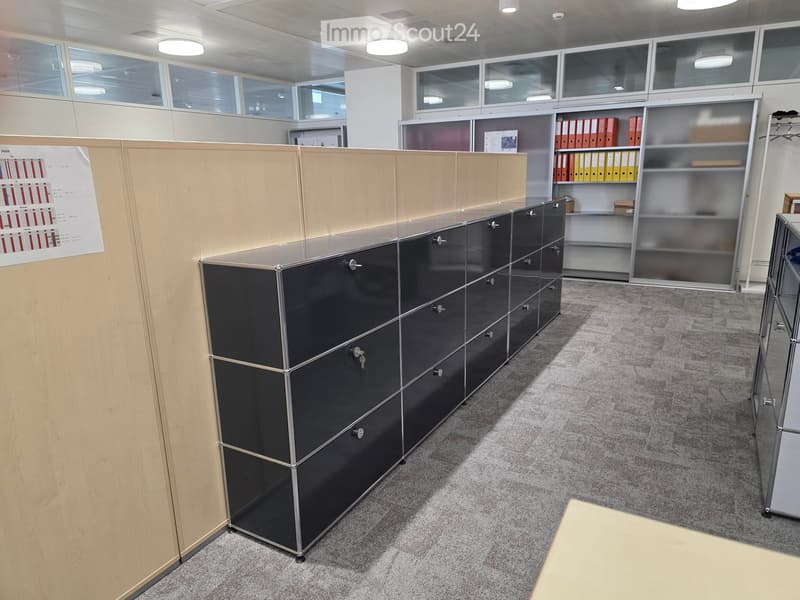 Modern office space with grey carpet, glass partitions, black metal cabinets, and shelves holding binders.