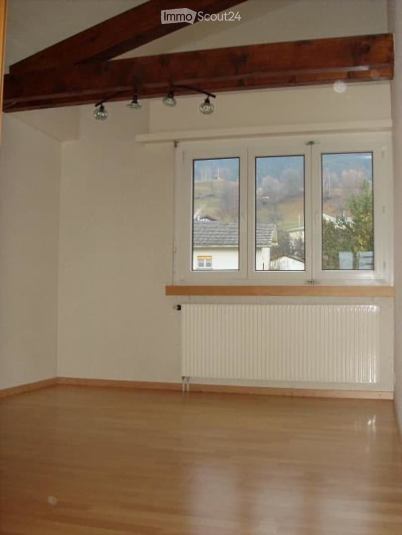Empty room with wooden flooring, white walls, large windows, and exposed wooden beams on the ceiling.