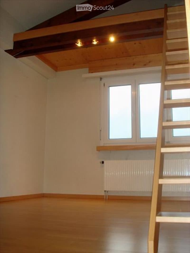 Empty room with wooden flooring, white walls, large windows, and a wooden staircase leading to an attic space.