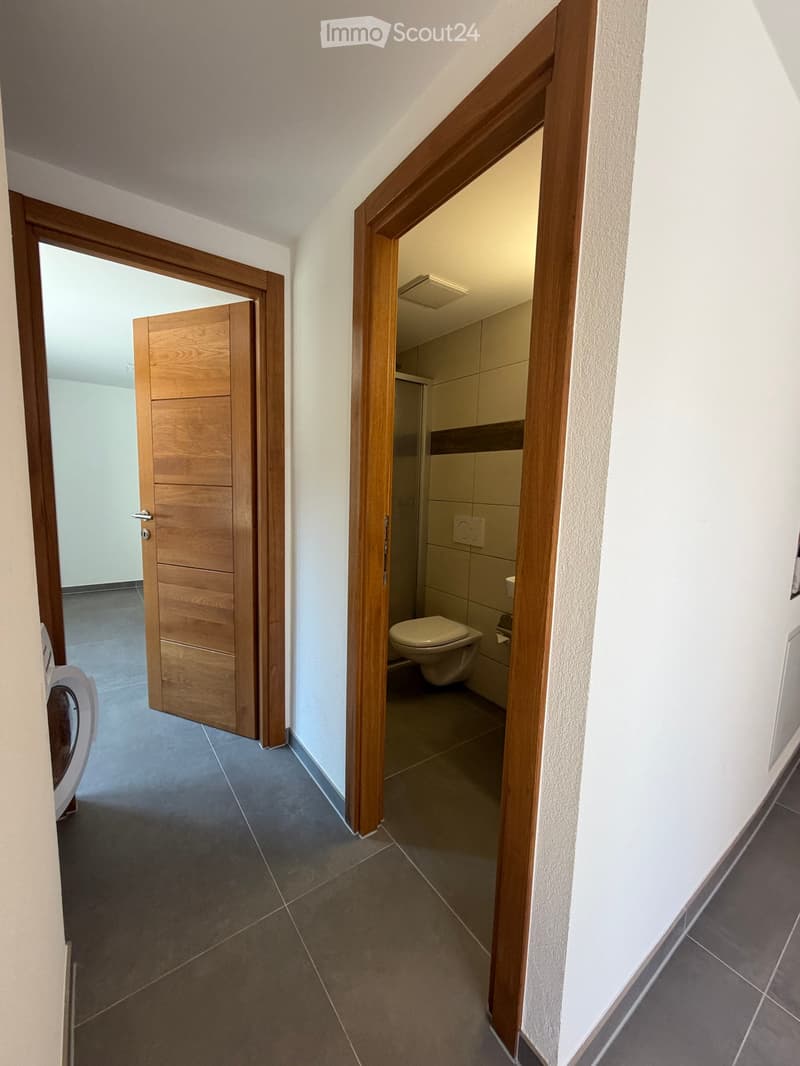 A narrow hallway with tiled flooring and white walls, featuring two wooden doors leading to different rooms, including a bathroom with a toilet and shower area visible through an open door.