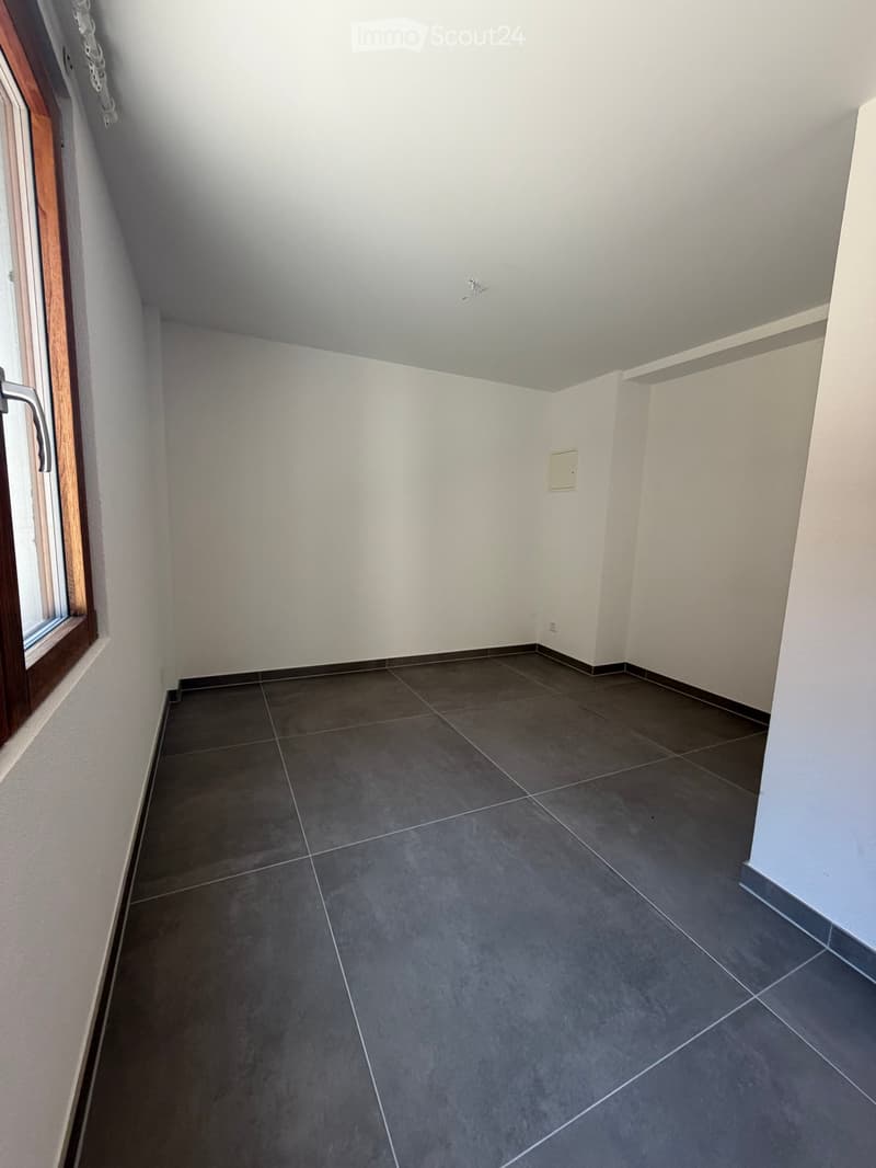 Empty room with grey tiled floor, white walls, and a window with wooden frame on the left side.