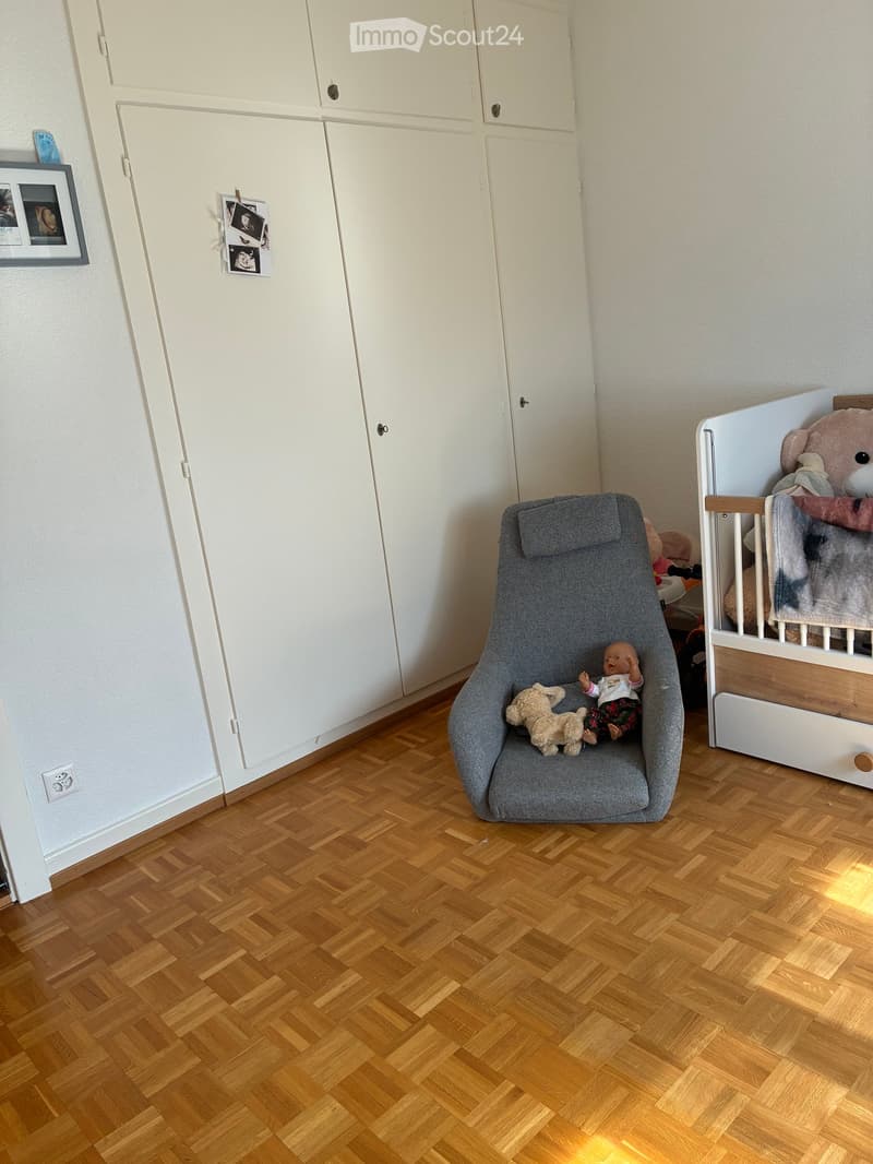 A well-lit children's bedroom with wooden flooring, a grey baby chair, a white wooden cradle, and a wardrobe with framed photos on the wall.