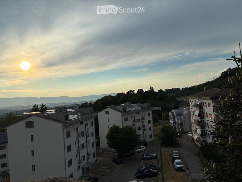View of a residential area with multiple apartment buildings, parked cars, and a scenic backdrop of hills and a setting sun.