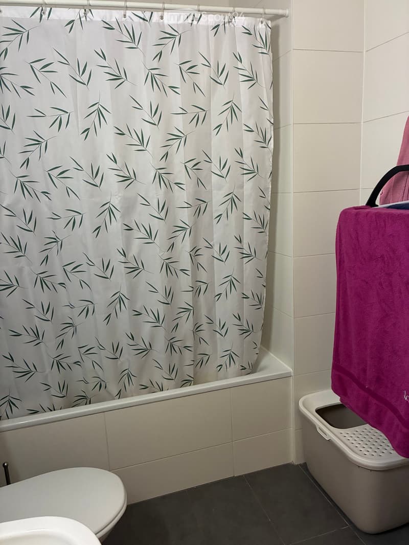 Bathroom with a bathtub, toilet, and a white shower curtain with green leaf patterns. A pink towel is draped over the edge of the bathtub.