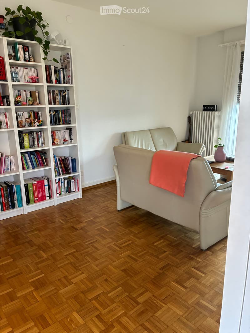 Living room with a cream sofa, a bookshelf filled with books and decorative items, a small table with a plant, and a radiator near the window.