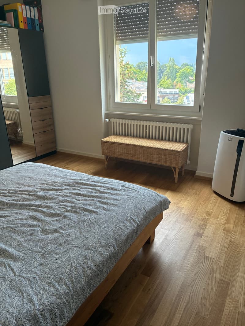 Bright bedroom with wooden flooring, large windows offering a view of greenery, a neatly made bed, a wicker bench, a radiator, and an air conditioner unit.