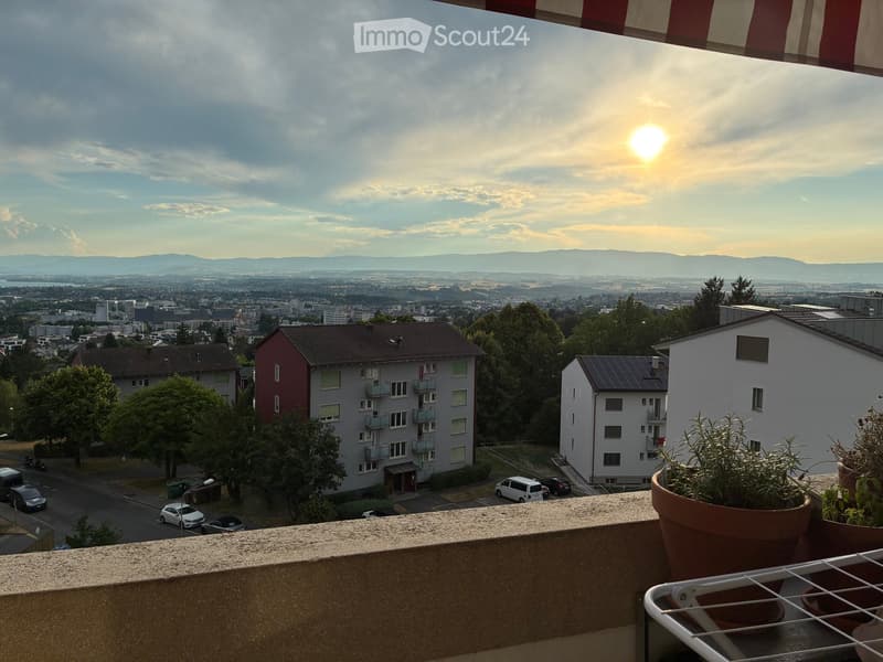 Balcony with a view of a cityscape, including buildings, parked cars, and distant mountains under a partly cloudy sky.