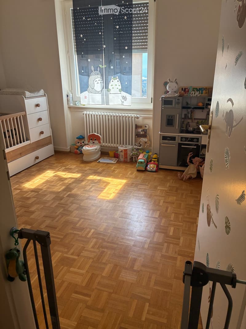A well-lit children's room with wooden flooring, a crib, microwave, oven, radiator, and various toys scattered around.