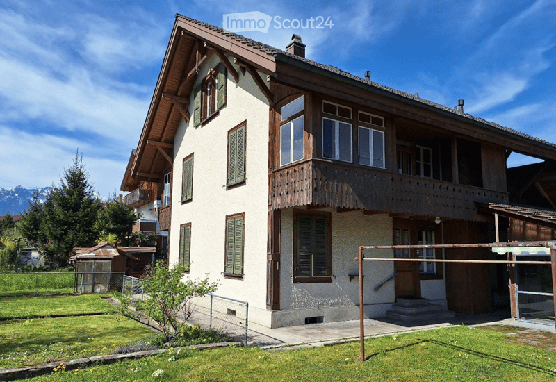 Two-story house with white walls, green shutters, wooden balcony, and mountain view.
