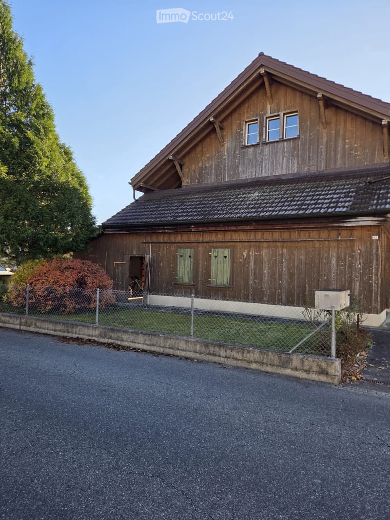 Two-story wooden house with a steeply pitched roof, multiple windows, and a fenced front yard with a small garden and a bicycle.