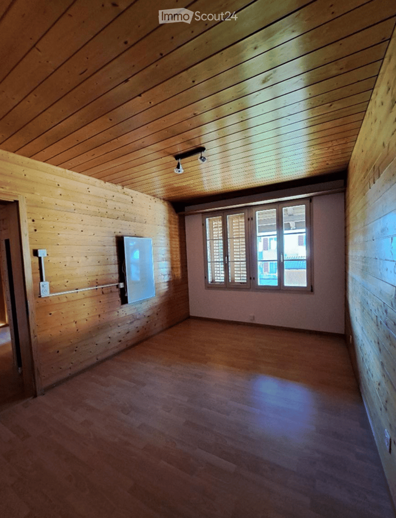 Empty room with wooden walls and floor, large window with shutters, mounted TV on wall, and recessed lighting.