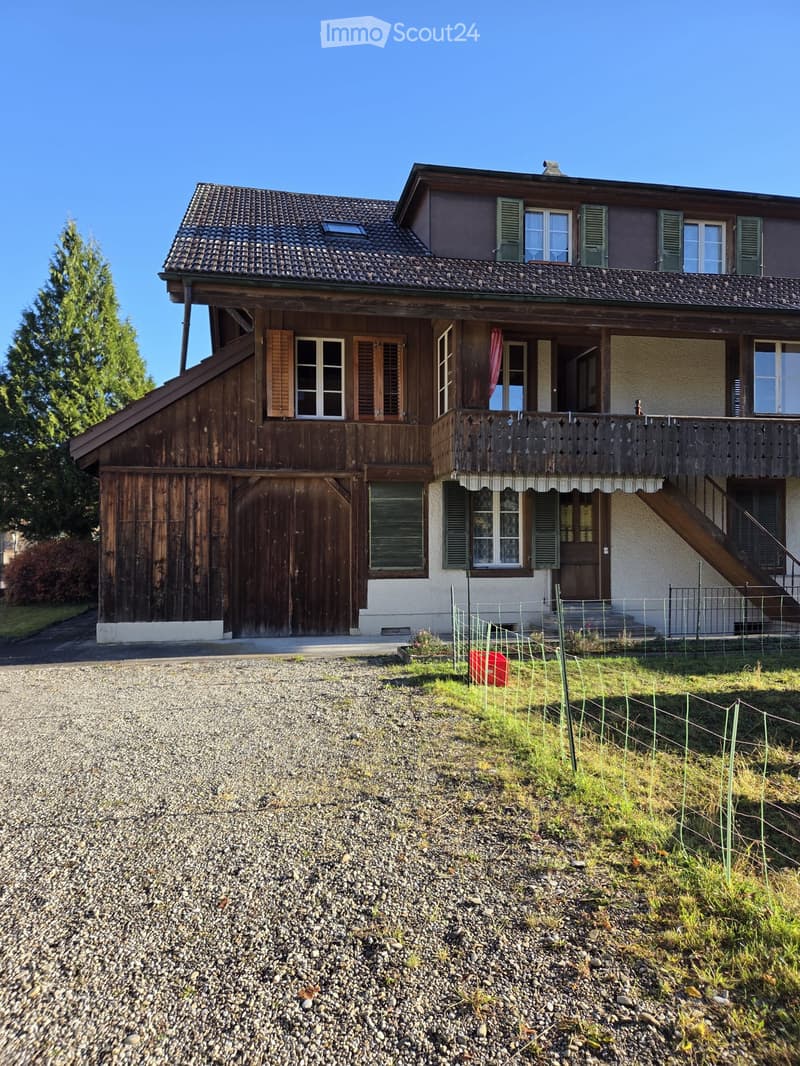 Two-story house with wooden balcony, multiple windows, and a garage. The house has a tiled roof and is surrounded by a gravel driveway and a small fenced garden area.