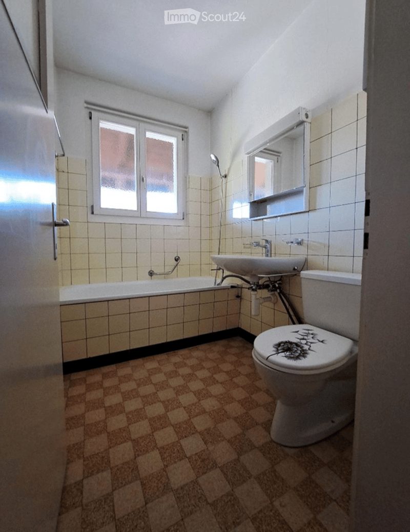 Bathroom with beige tiled walls, a bathtub, a toilet with a decorative sticker, a sink, and a mirror above it. The floor is covered with brown checkered tiles.