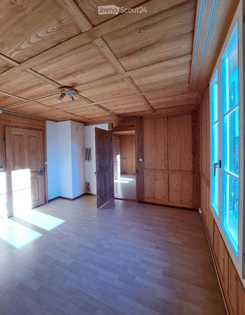Empty room with wooden flooring and exposed wooden ceiling beams. Natural light streams in from large windows, and there are several wooden doors leading to other spaces.