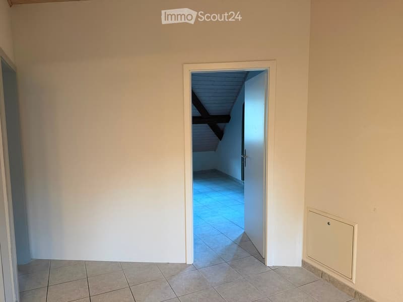 Empty hallway with tiled floor, white walls, and an open doorway leading to another room with a slanted ceiling and tiled floor.
