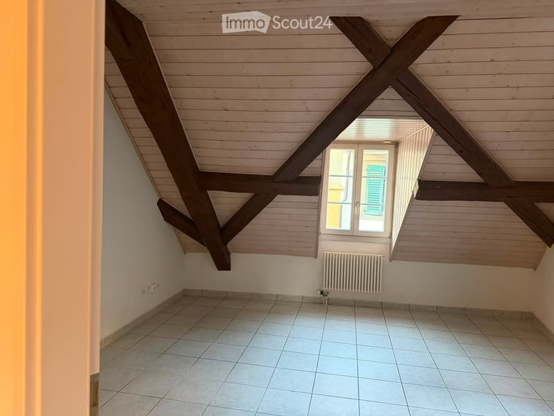 Empty attic room with exposed wooden beams, tiled floor, and a single window with a radiator below.