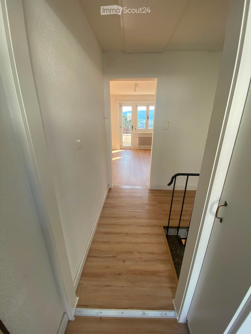 A narrow hallway with wooden flooring, white walls, and a staircase leading upwards. The hallway has a handrail on one side and a door at the end leading to another room with large windows.