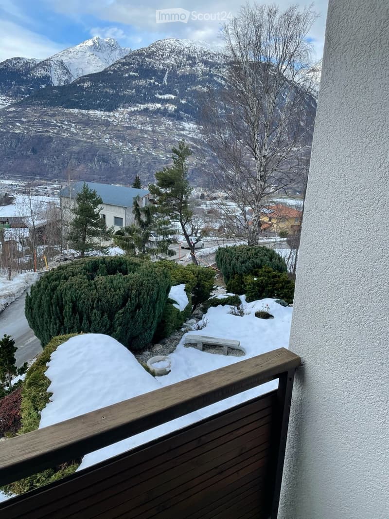 balcony with wooden railing, snow-covered ground, bench, shrubs, trees, distant house, mountains
