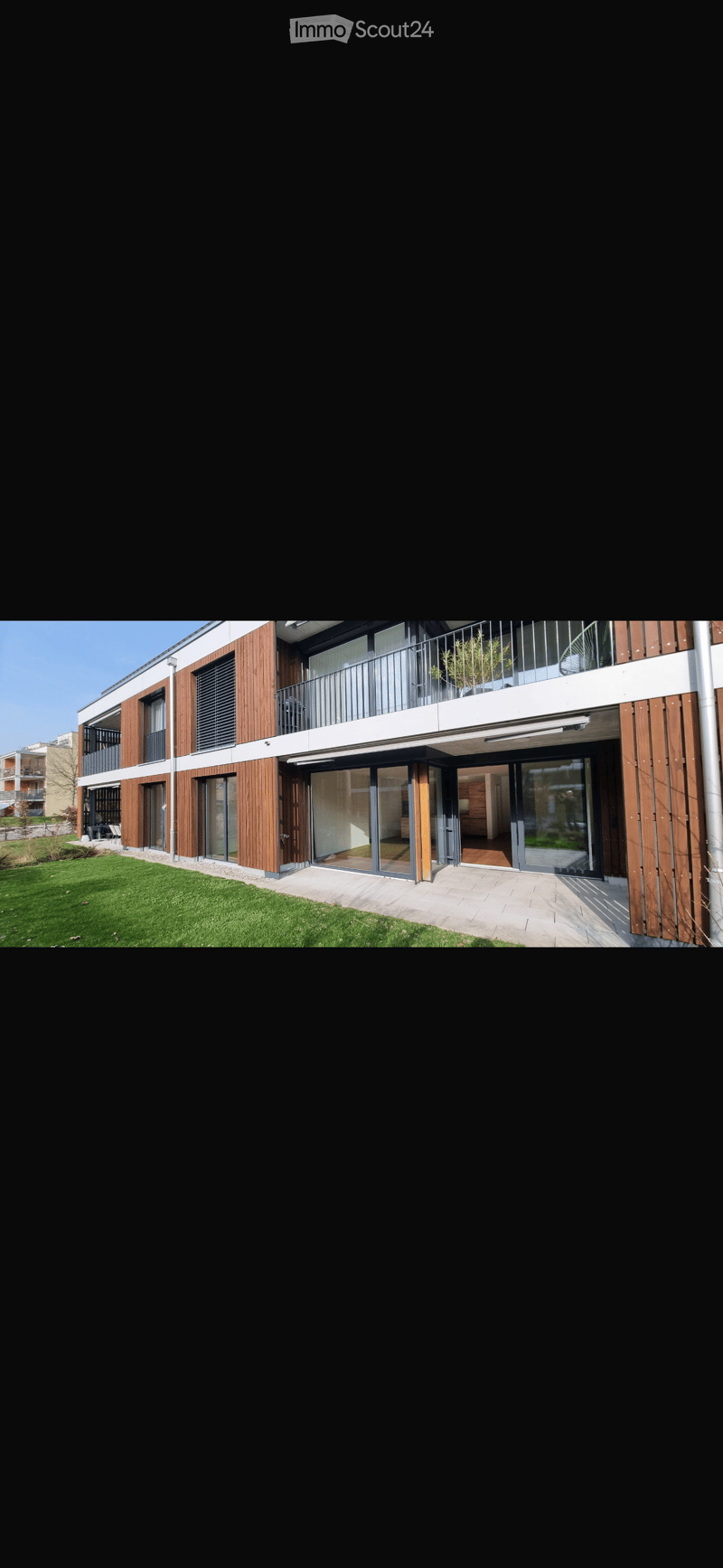 Modern multi-story residential building with wooden paneling, large windows, and balconies. The entrance features a glass door leading to an interior space visible through the glass.