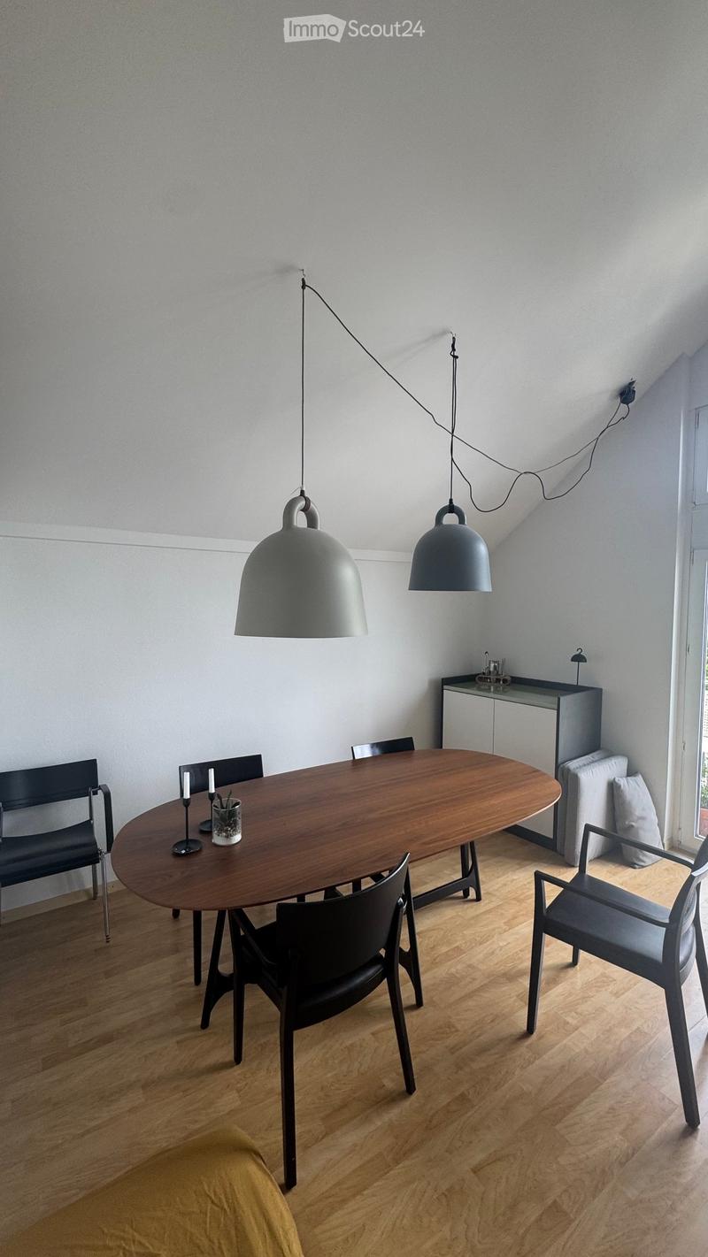 Modern dining area with a round wooden table, black chairs, pendant lamps, and a small cabinet with a sofa nearby.