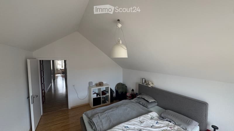 A cozy attic bedroom with wooden flooring, a large bed with grey bedding, a nightstand, a hanging lamp, and a small shelf with books and decorative items.