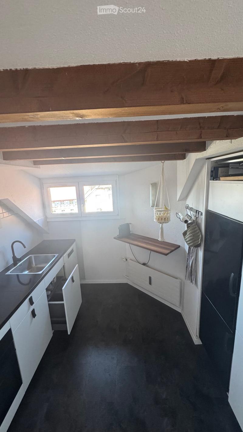 Modern kitchen with white cabinets, black countertop, stainless steel sink, and a wooden shelf with a hanging basket. The space features large windows and a dark floor.