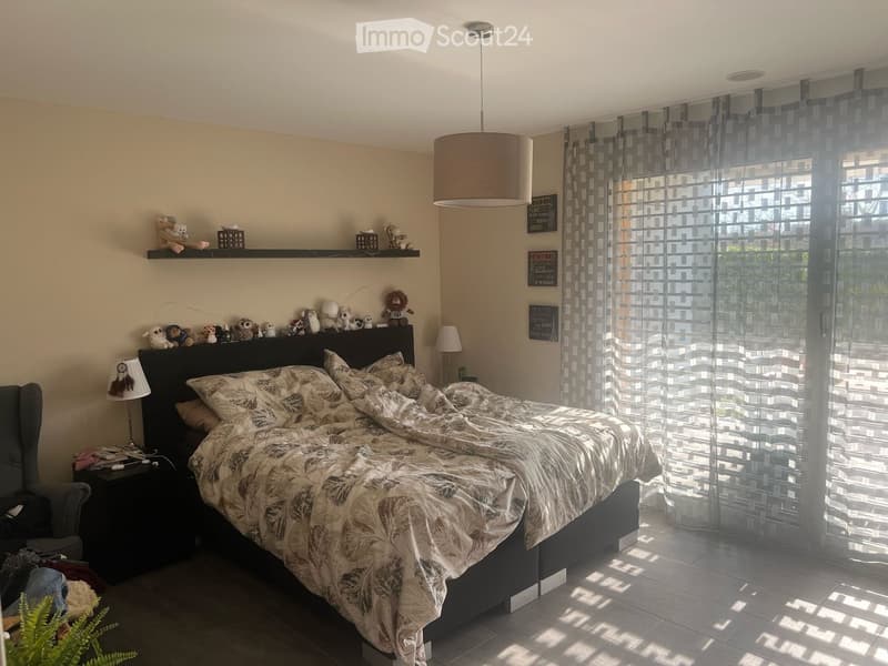 A well-lit bedroom with a large bed, nightstand, and decorative shelf holding stuffed animals. The room features modern curtains and a sleek lamp.